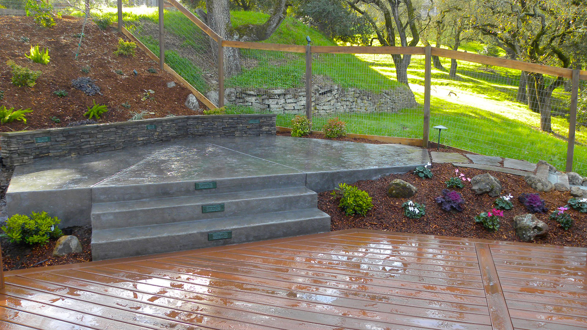 Stamped concrete patio, framed by a ledgestone seat wall, steps down to and contrasts with a Fiberon synthetic deck